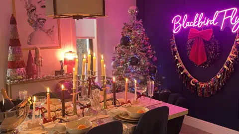 IWC Media Festive dining room with a decorated Christmas tree, a neon sign reading “Blackbird Fly,” a table set with candles and holiday décor, and colourful garlands.