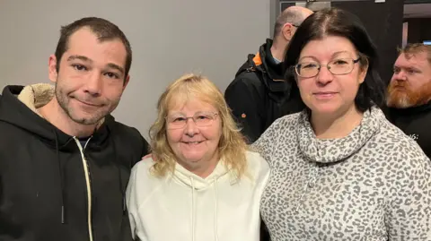 Three local people, two women one man, with arms around each other smiling. Both women are wearing glasses and jumpers. The man is wearing a black hoodie.
