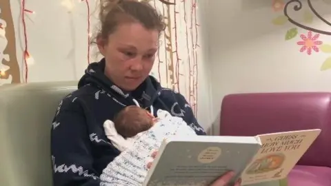 A woman in a blue top is holding a baby in front of her and holding a book in her other hand. She is sitting on a green chair with a purple sofa in the background and patterns and lights on the walls.