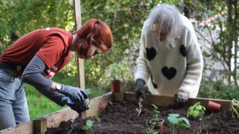 Avon Wildlife Trust Two women planting young plants in a garden in Easton in Bristol. The soil is dark and the plants they are placing in the planter have light green leaves