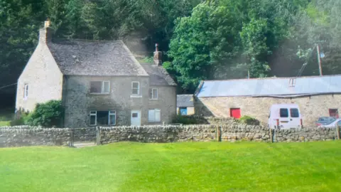 Plankey Mill farmhouse is a stone built cottage with a smaller extension and two chimney pots and outbuildings. The cottage is encircled by a stone wall with a white van parked outside.