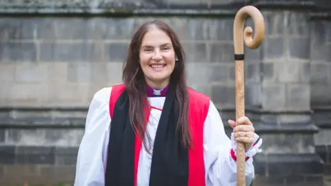 The Right Reverend Sophie Jelley, with long hair, is standing outside a building. She is facing the camera smiling and wearing clerical wear, including a red robe and collar. 
