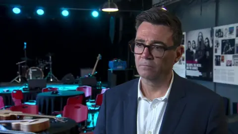 Andy Burnham. He has dark hair, wearing glasses, a navy blazer and white shirt.