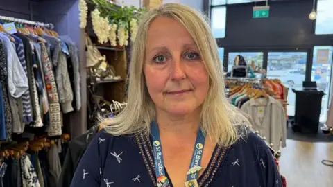 Grace McGrory/BBC A woman with blonde shoulder-length hair. She is wearing a St Barnabas lanyard and a dark blue dress with a white dragonflies pattern.