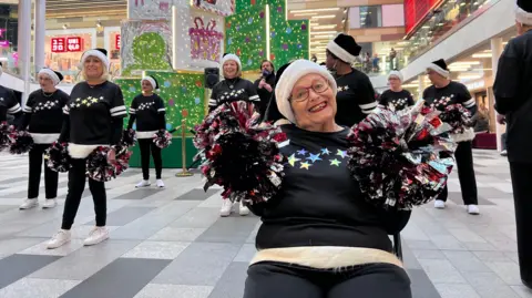 Lily-May Symonds/BBC A group of people wearing matching black sweaters with star designs and festive hats perform a cheer routine in a shopping centre. In the foreground is a woman sitting down with a big grin on her face.