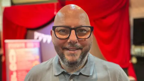 Man wearing glasses and a polo top in front of a stage