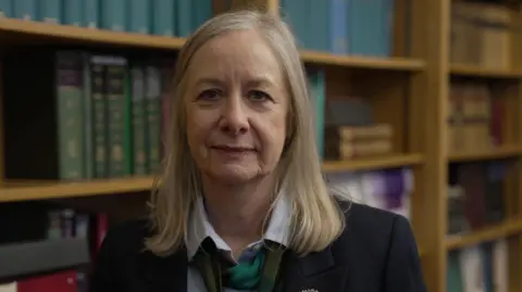 Ashley Edwards KC, a blonde-haired lady wearing a dark suit jacker, grey shirt and a loosely knotted tie, stands in front of a full bookshelf, presumably in her legal office.