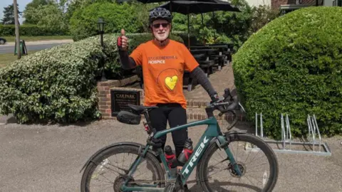A man in an orange t-shirt and long black sleeve shirt is standing by a bicycle. He is wearing a helmet and a pair of sunglasses.