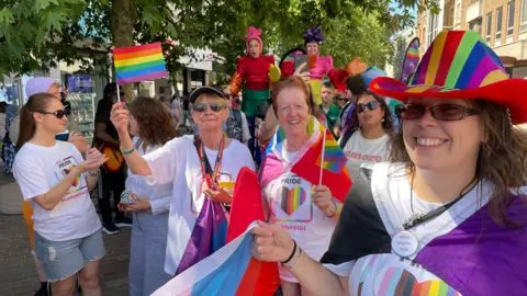 Sam Read/BBC A group of women holding small pride flags on white sticks. They also have flags wrapped around their shoulders. They are walking through the town as part of the parade and crowds of people can also be seen behind them.