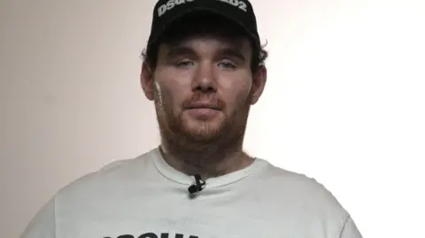 Luke looks into the camera with a serious expression. He is wearing a dark baseball cap and a white t-shirt. He has a short reddish-brown beard