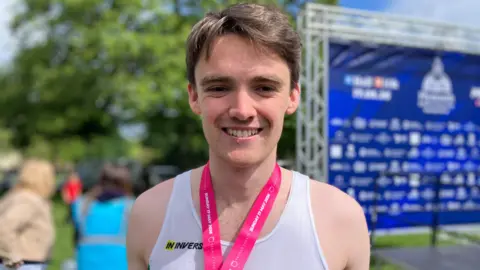 A man with brown hair smiling at the camera. He is wearing a white t-shirt and pink lanyard. The background is slightly blurred. There is a stage to the right and people in the background to the left. 