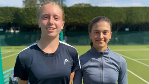 Daniella Britton (left) and Grace Watson stand between two tennis courts, smiling at the camera. Daniella has ginger hair and dental braces and wears a black T-shirt. Grace, who is standing slightly further back and appears to be the shorter of the two, has dark brown hair and wears a grey, zipped sports top.