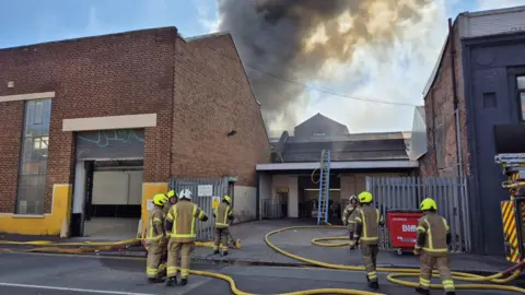 Seven firefighters dressed in brown firefighting uniforms with yellow helmets work at the scene of a fire. Smoke is billowing into the sky behind a building in the background. Yellow hoses from a fire truck on the right can be seen snaking along the ground and a ladder has been placed against a roof in the background.