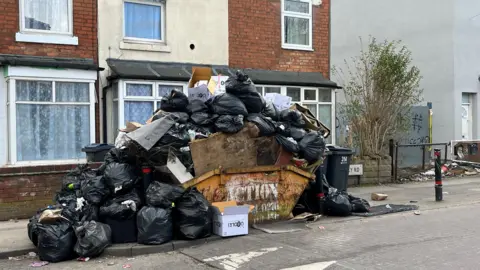 A pile of black bin bags and rubbish piled high on top of a skip that is on the pavement of a residential street.