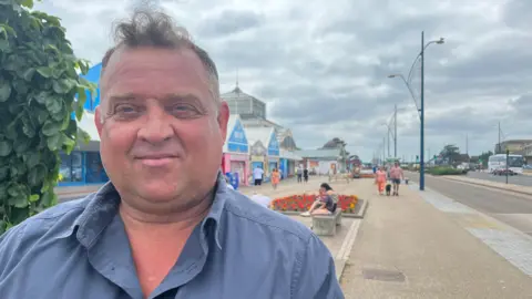 Andrew Turner/BBC Asa Morrison, standing outside on the seafront. He is standing on the left of the frame and is looking directly at the camera and smiling. He is wearing a grey shirt.