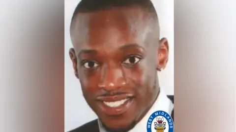 West Midlands Police A man with cropped black hair and a trimmed moustache and beard is wearing a white collared shirt and black jacket. He is looking at the camera, in front of a white background.