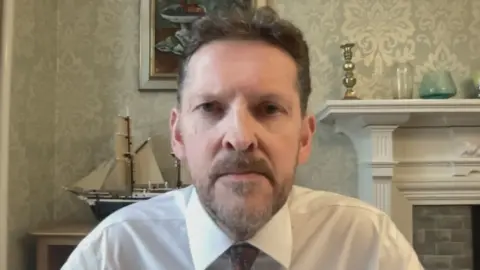 "Dr Ian Kennedy wearing a white shirt and patterned tie, facing the camera in a room with patterned wallpaper, a model sailing ship, framed artwork and a white fireplace mantel in the background."