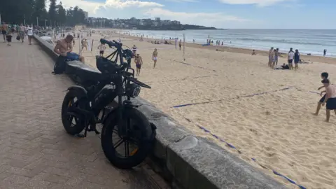Helen Livingstone A fatbike on a promenade in front of a beach where people are playing volleyball