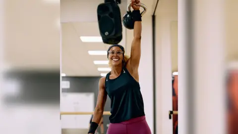 A person wearing a black vest top and purple leggings holds a kettle bell in the air and smiles. In the background you can see a gym studio with a speaker hanging from the ceiling.