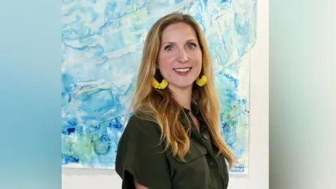 Abi Spendlove Abi Spendlove smiling at the camera in front of one of her artworks that has shades of blue representing water. She has golden long hair and wears large yellow earrings and an olive green short sleeved shirt dress.