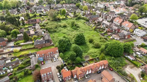 Tom Watson A bird's-eye drone shot of a green space in Driffield, which includes large bushes and trees. It is surrounded by residential houses and gardens. 