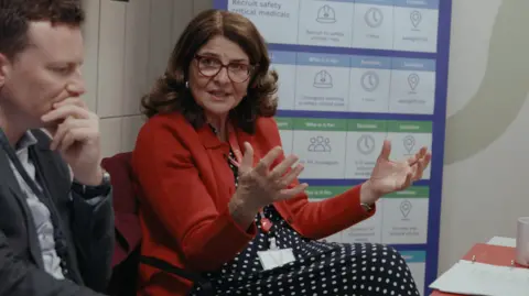 Widnell William/DWP A woman in a red coat and black and white spotty dress speaks passionately to a group at a table. 