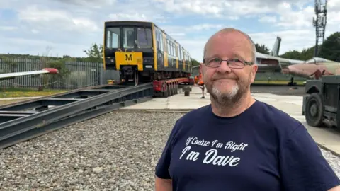 David Charles has short grey hair and glasses. He is wearing a navy blue t-shirt reading Of Course I'm right I'm Dave. And old Metro train is behind him.