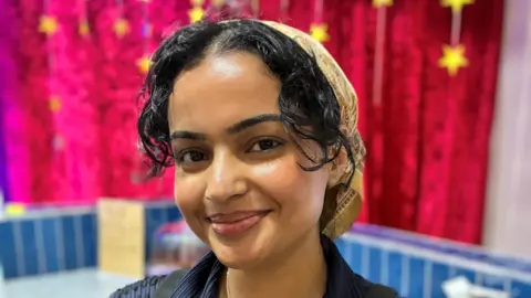 A woman with her hair tied back smiles at the camera. She has dark hair and is wearing a blue blouse. 
