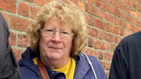 A woman with curly blond hair and wearing glasses, a yellow jumper and a blue coat, is standing in front of a brick wall.