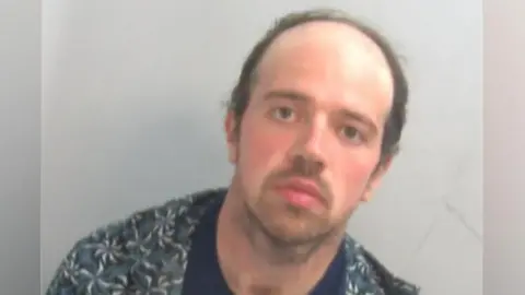 A police custody mugshot of Luke D'Wit, who is wearing an unbuttoned shirt with a T-shirt underneath. He has  short dark hair and a blank expression on his face.