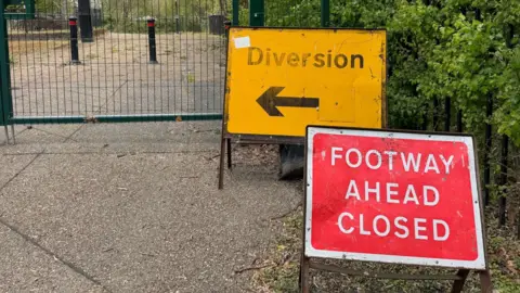A red road sign with the words "FOOTWAY AHEAD CLOSED" in white lettering in front of a yellow road sign with the word "Diversion" above a black arrow. Both signs are on a pavement in front of a bridge which is fenced off to the public.