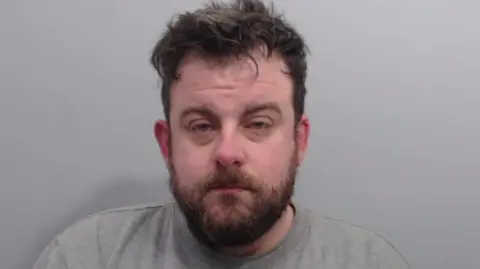 A man looks into the camera. He has a dark beard and dark, dishevelled hair. He is wearing a grey, round neck top and is standing against a grey background.