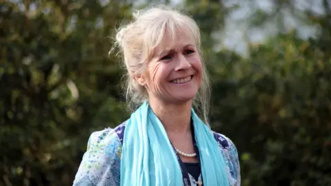 Brunel Academies Trust A woman with blonde hair scraped back into a bun with a fringe, smiling, and wearing a blue, white and lime green patterned blouse, a baby blue scarf around her neck and a pearl necklace. The background is slightly blurred with trees, leaves and grey sky.