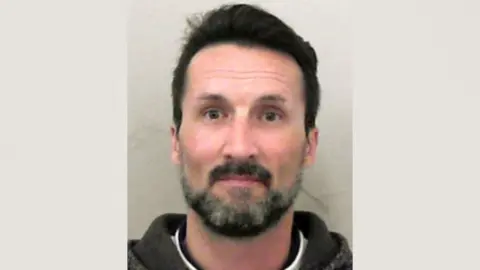 A police mugshot of Mark Acklom. He is wearing a white T-shirt underneath a black hoodie. He has short dark hair and a dark beard with grey patches. 