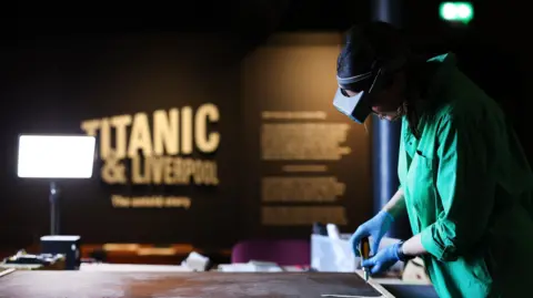 Gareth Jones/National Museums Liverpool A female worker works on preserving a painting rolled out on a table in front of her. She wears goggles, a green overall and blue latex gloves. The sign Titanic and Liverpool can be seen behind.