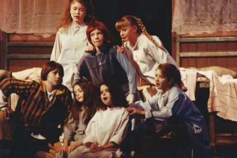 Phoenix Theatre A young Laura Main, in a bright red "Annie" wig, on stage with the cast of Annie in a youth production. The scene is set in the dorm of an orphanage