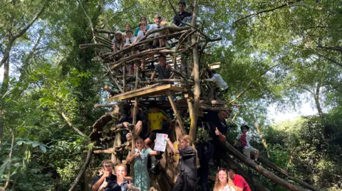 About a dozen children play inside, on top of and around a large treehouse built out of logs and wood planks. It is inside a canopy of trees.