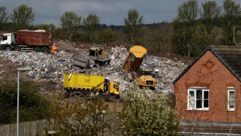 A landfill site with several lorries working on the site. A red brick house that backs onto the quarry with only a short wooden fence separating them. 