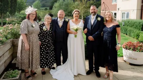 Dormy Care Communities The couple and their family, including Rachel's mother, Ruth, smiling outside in the garden of the carehome. The woman far left is wearing a leopard print dress with a large white hat, she has dark reddy-brown hair. Next to her is an older woman in a black and white dress. To the right of her is a balding man with a blue three piece suit and cream tie and flower. To the right of him is a woman in a white wedding dress holding a bunch of white flowers. Next to him is a man who has short grey-black hair and a blue three piece dark blue suit, he also has a cream tie and flower. And finally to his right is a woman with shoulder-length blonde hair and a blue off the shoulder flowery dress. They are stood in the garden and you can see trees, grass and flowers behind them. To the right is a cream and right bricked building. 