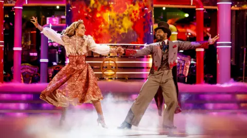The dance partners dressed in Victorian-style fancy dress, dancing on in a ballroom with pink lights and smoke around them. JB wears a brown suit with a waistcoat and top hat, while Amy wears a dress with a white blouse on top and an embroidered peach-coloured skirt. 