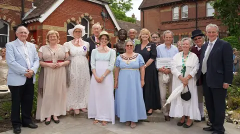 East Hampshire District Council A group photo of guests attending the statue's unveiling ceremony at Alton's Regency Garden, including the author's fifth great niece Caroline Knight and town mayor Annette Eyre. Some guests are wearing Regency clothes. The group is surrounding the statue. It is a cloudy day.