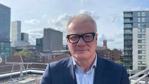 A man wearing thick black glasses with light blond to grey hair stands on a balcony with Birmingham's skyline behind him.