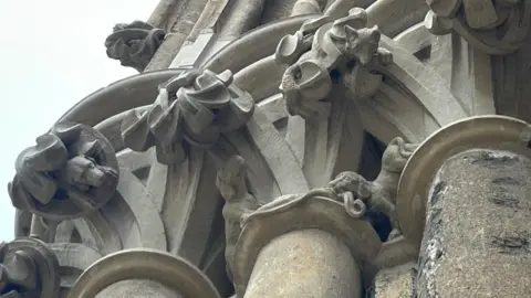 An intricate stone carving of a hunting scene situated around the pillar of a cathedral. Two dogs can be seen at the bottom, with one looking up towards a fox hiding higher up. Another dog can be seen to the left of the pillar.