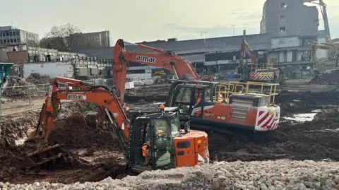 Three dark orange excavators are working in footings of the development. in the background is the remnants of the old NTL building.