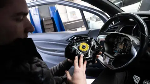 Getty Images A man sat in a car's driving seat takes apart a steering wheel to replace a faulty airbag system.