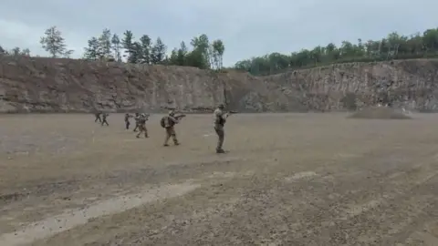 An image showing multiple men in the distance wearing combat military gear and holding firearms. They are walking in an empty field overlooking a rocky hill
