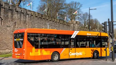 An orange single decker bus in traffic driving past Cardiff Castle.
