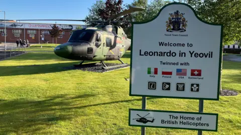 Leonardo Helicopters site in Yeovil. A helicopter can be seen on the lawn and a sign reading "Welcome to Leonardo in Yeovil" is in the foreground of the picture.