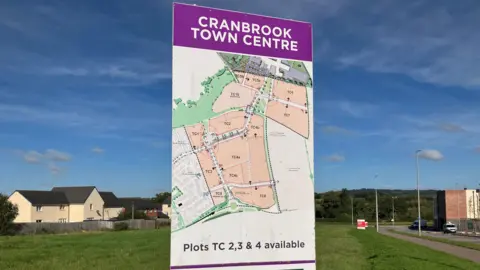 A sign for Cranbrook Town Centre on a large patch of grass showing different plots in the area with houses and shop buildings in the background on a sunny day.