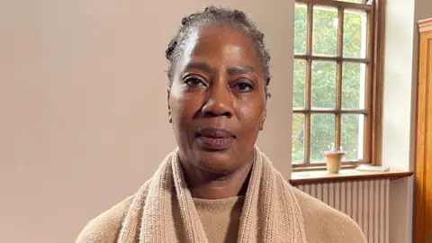 Sylvia Turay, looking directly to camera. She is sitting in front of a blank, magnolia wall, with a large sash window over her left shoulder. She is wearing a cream coloured top, overlayed with a large knitted cream scarf. She has black hair and dark brown eyes.